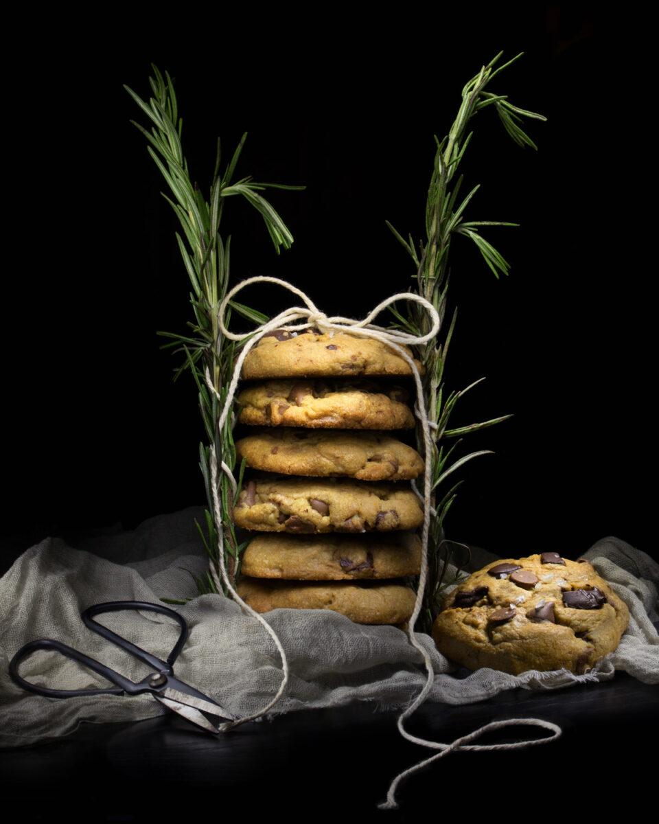 Still life photography of a stack of chocolate chips cookies with rosemary around it.